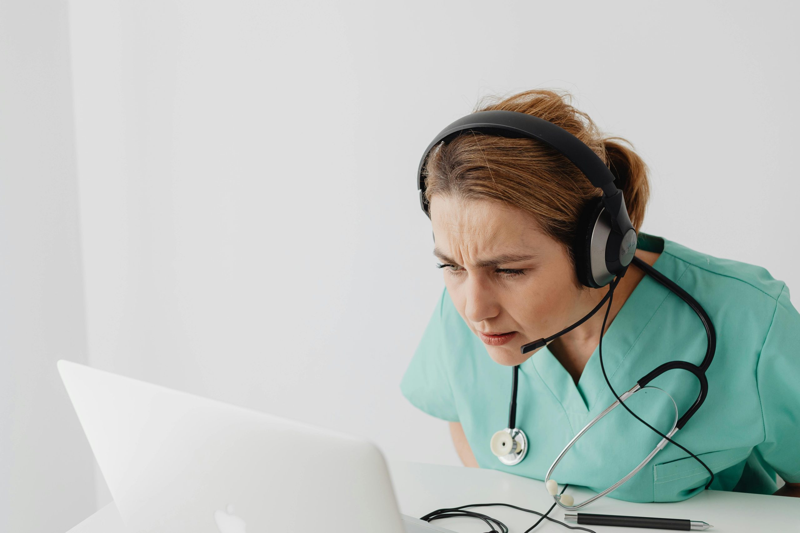 Female doctor wearing scrub suit and headset during an online video call with a laptop.